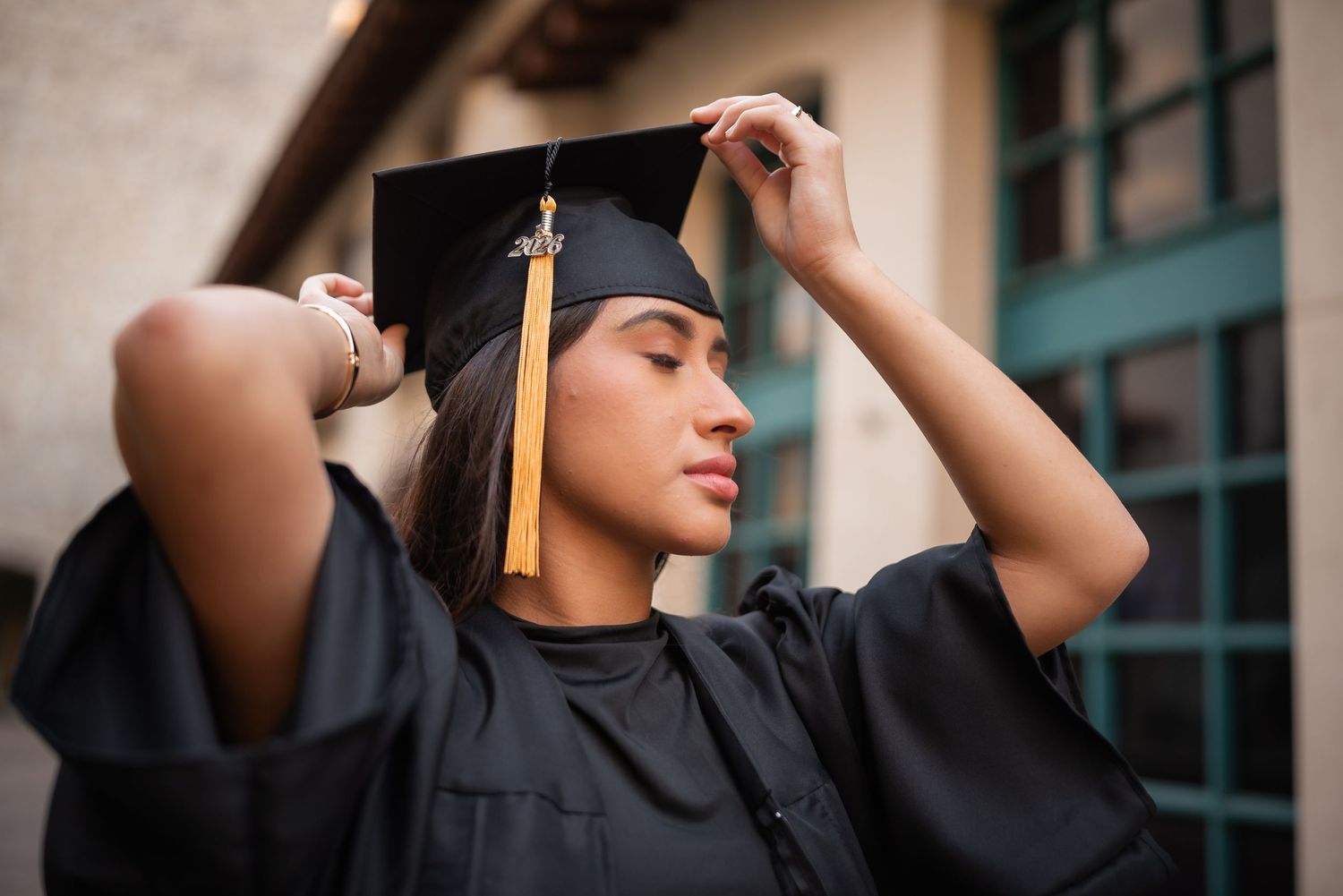 Young female graduate in black cap and gown adjusting her graduation cap with gold tassel outdoors near a campus building