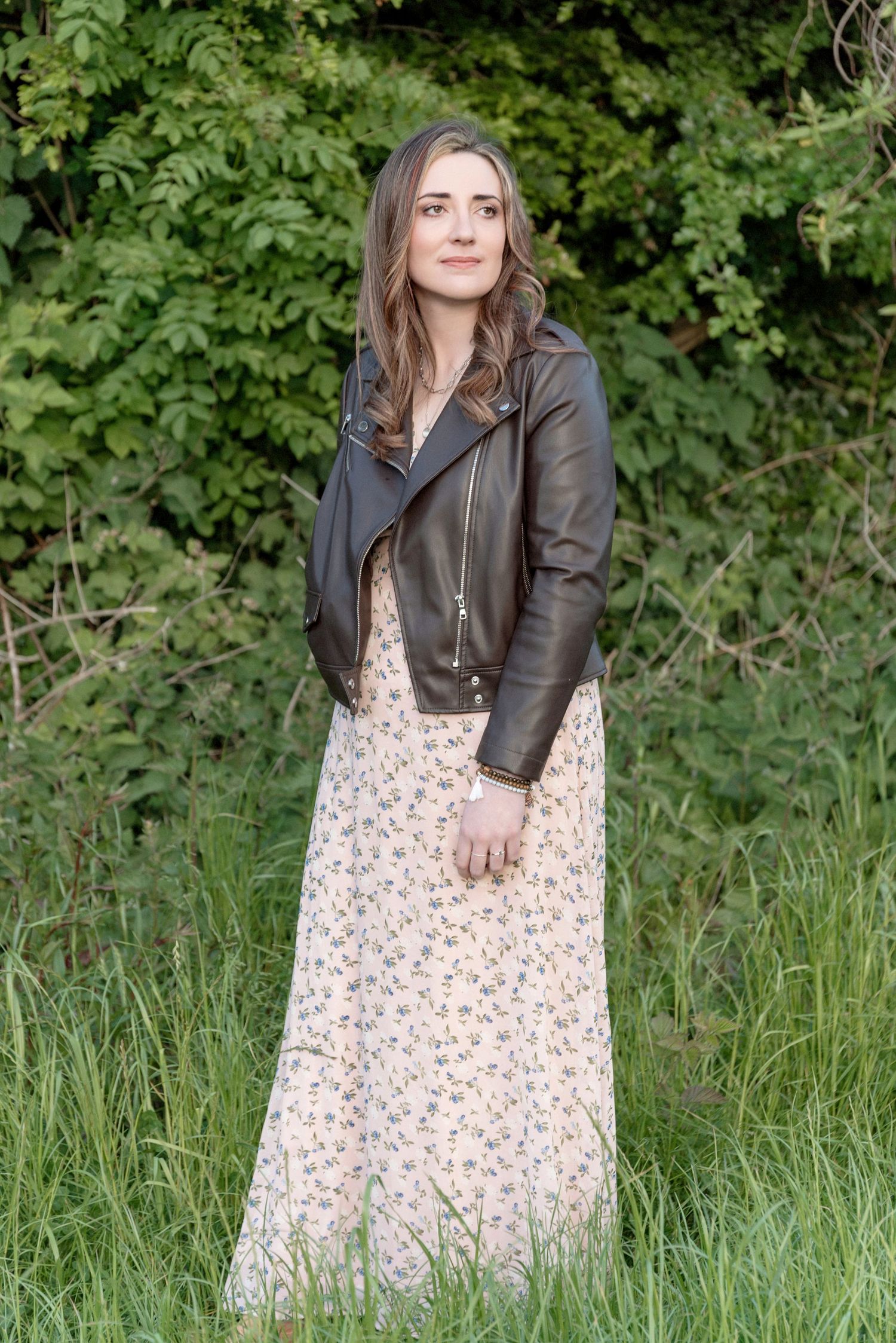 A model wearing a brown leather jacket and floral maxi dress poses against a backdrop of green foliage.