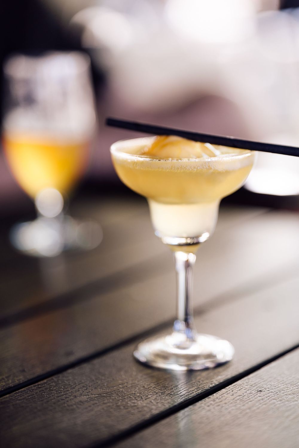 A frothy yellow cocktail in a margarita glass served on a dark wooden bar.