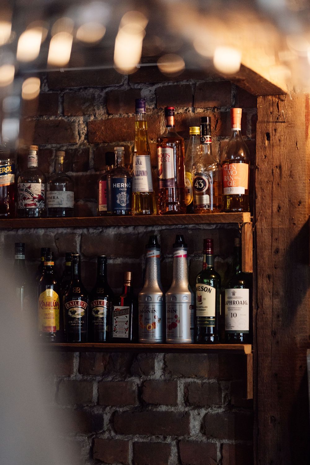 Rustic wooden shelves lined with assorted liquor bottles against a brick wall.
