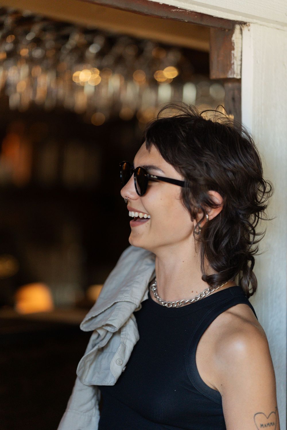Smiling woman with short dark hair wearing sunglasses, black tank top, and silver chain necklace, leaning against a wall.