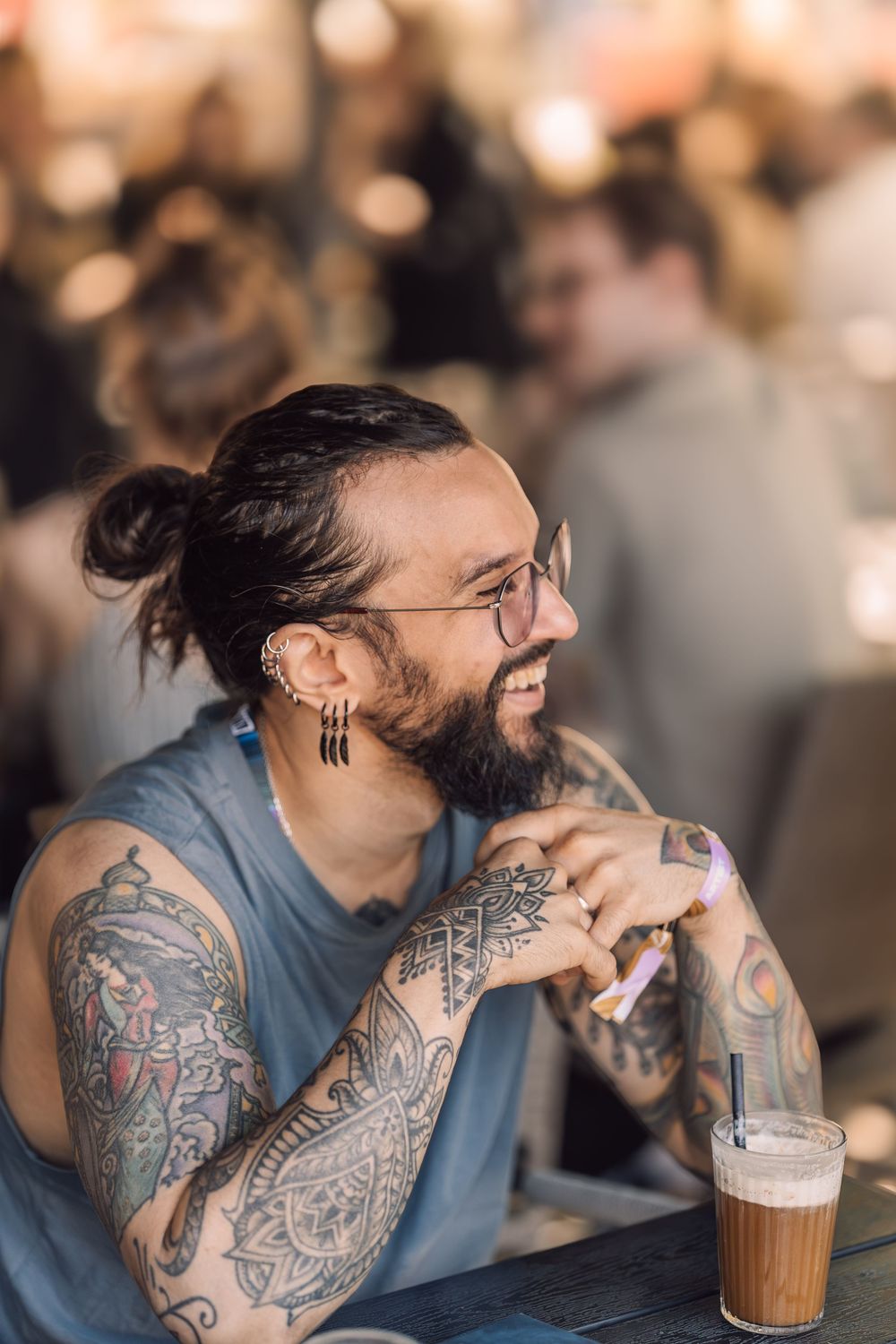 Smiling tattooed man with hair bun and glasses sits at a bar table with an iced coffee drink.