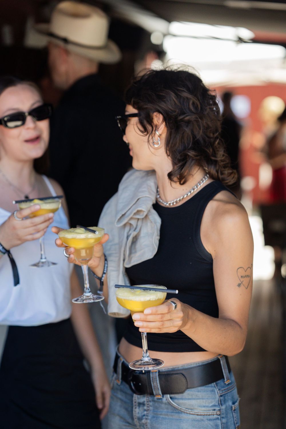 Two women wearing sunglasses enjoy yellow cocktails at an outdoor bar event.