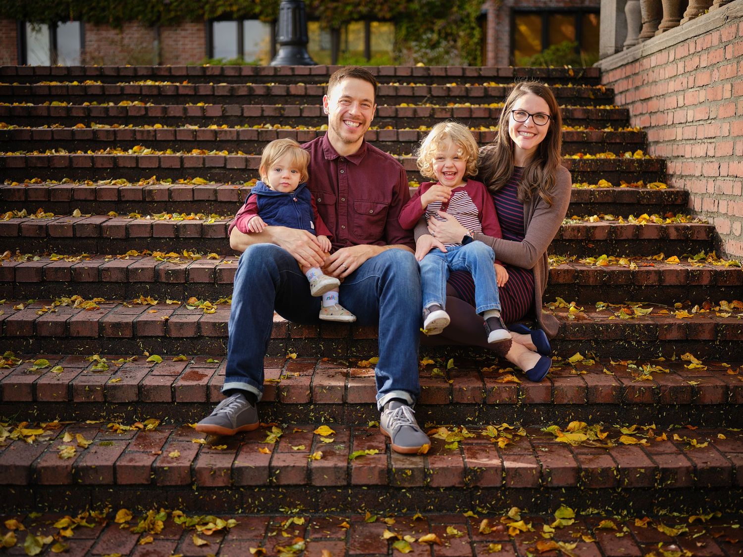 A family sits together on outdoor brick steps covered in colorful autumn leaves during a fall photo session.