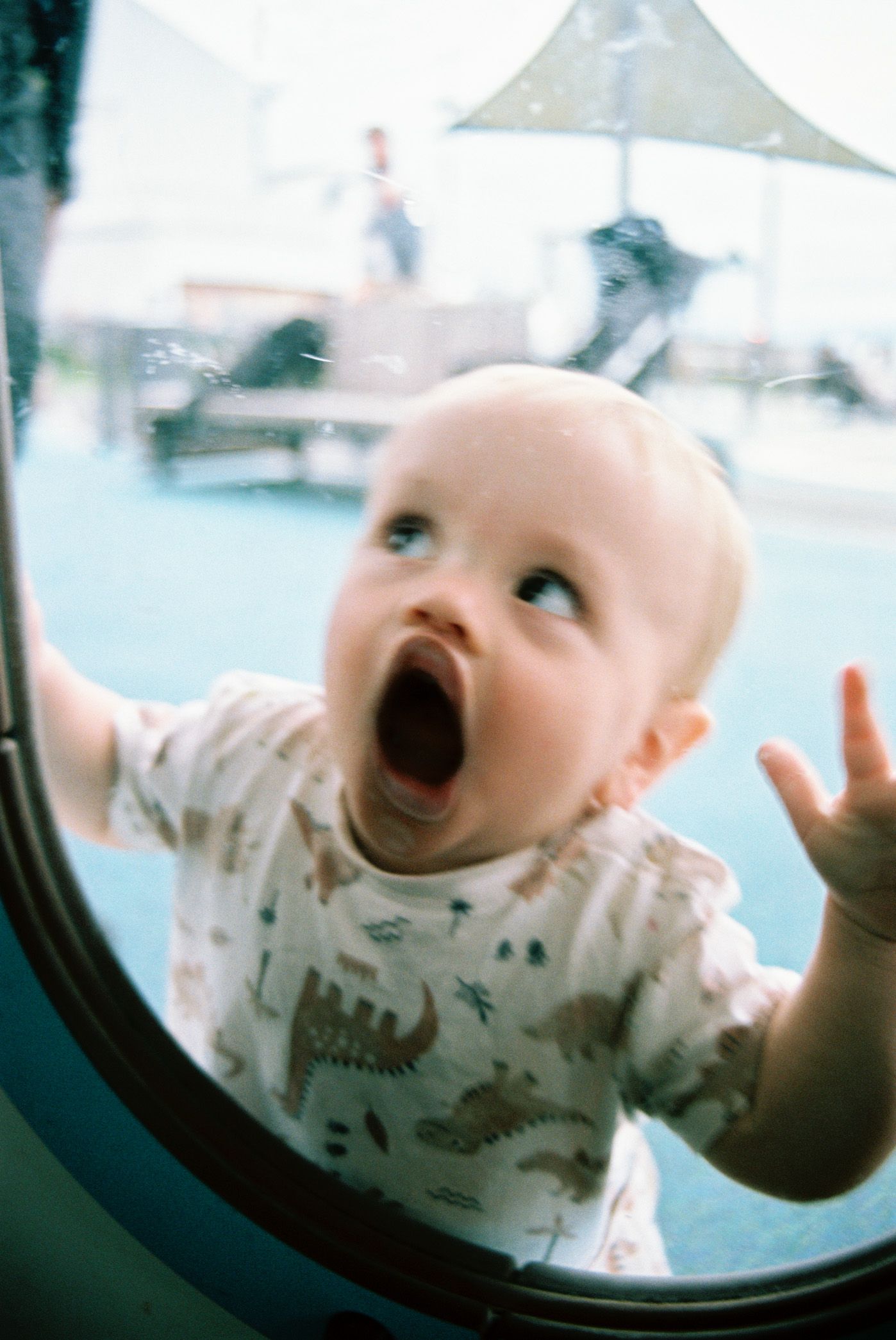 A small baby presses against a window with an excited open-mouthed expression while wearing a dinosaur print t-shirt.