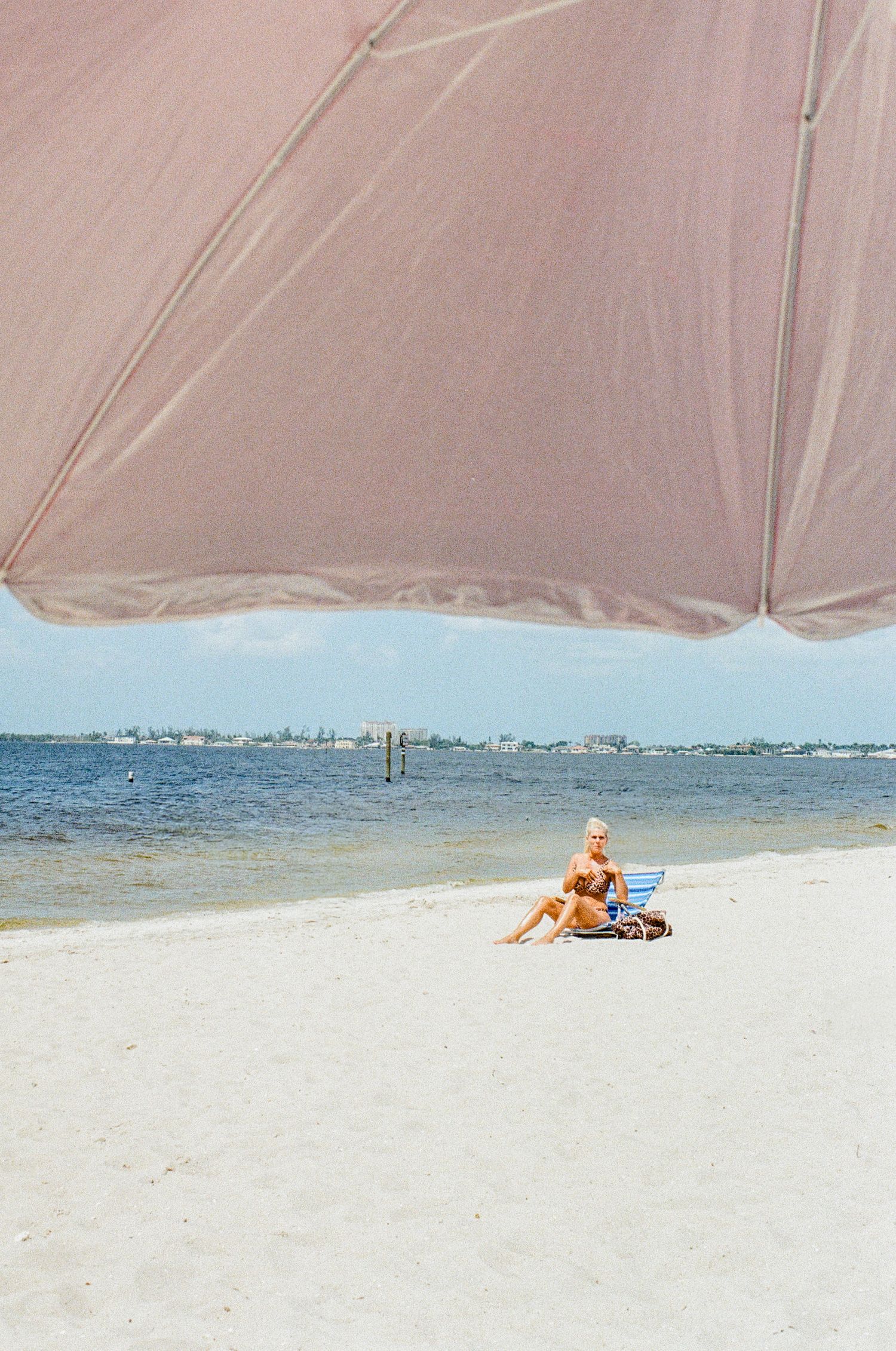 A beachgoer relaxes under a beige umbrella on a white sandy beach with blue ocean and distant buildings visible.