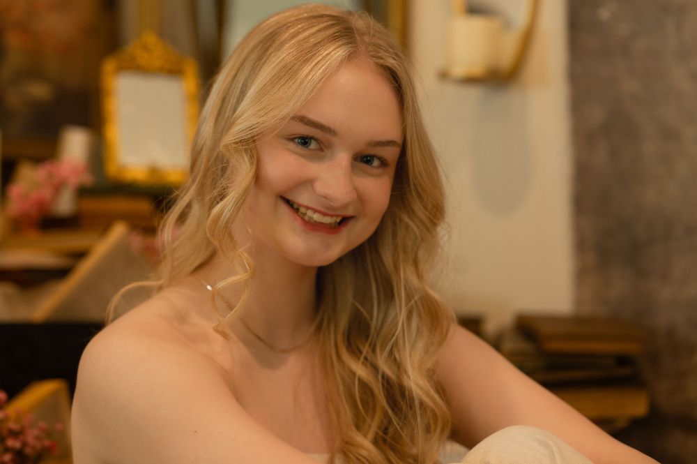 A smiling person with blonde wavy hair sits in a warm-lit indoor setting.