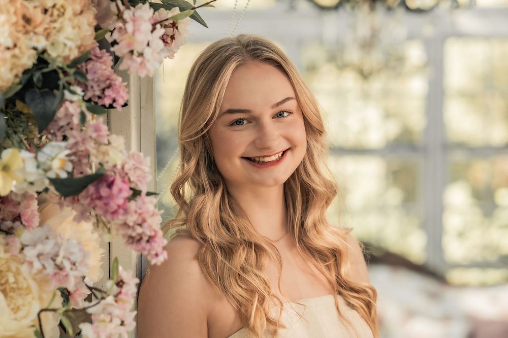 Portrait in a sunlit conservatory with pink floral arrangements and soft natural lighting.