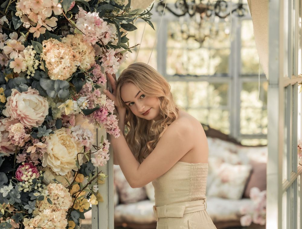 Elegant portrait in cream dress amid peach and pink floral arrangements in a conservatory setting.