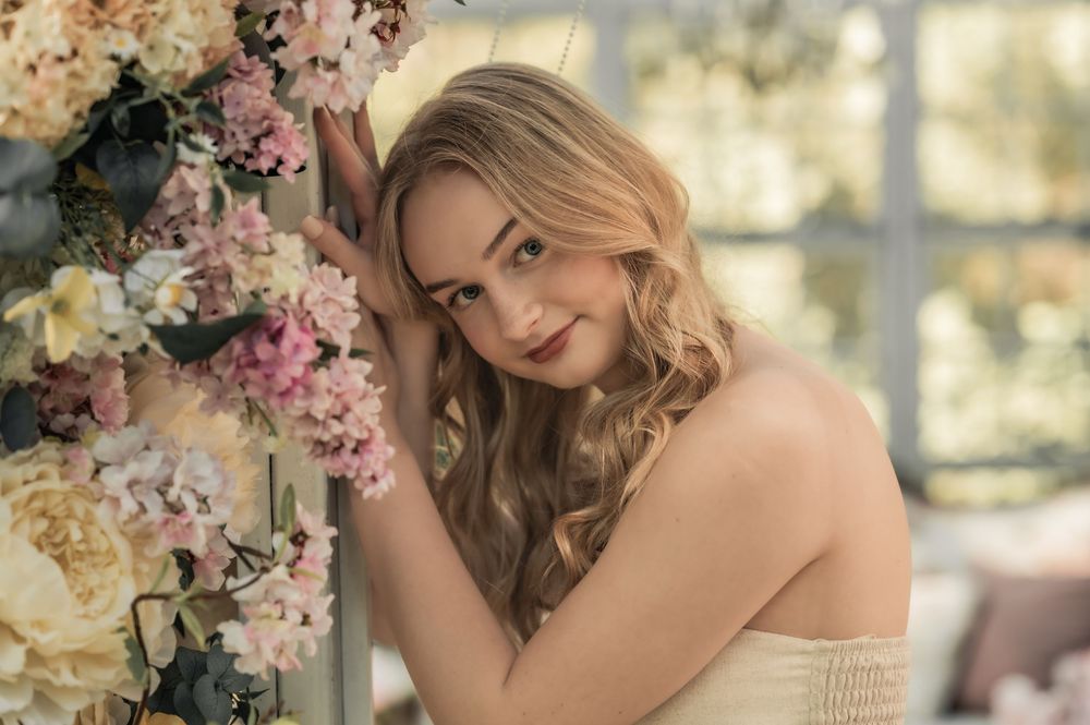 Person in off-shoulder dress poses next to floral arrangement with pink and cream colored blooms in romantic setting.