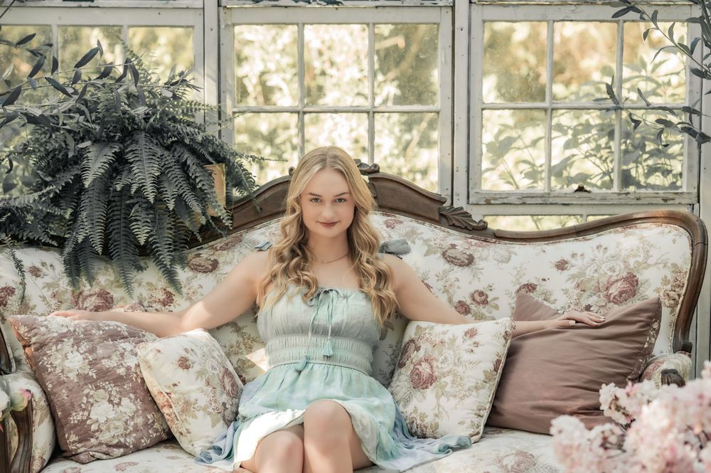 A mint green dress adorns someone resting elegantly on a vintage floral sofa with decorative ferns nearby.