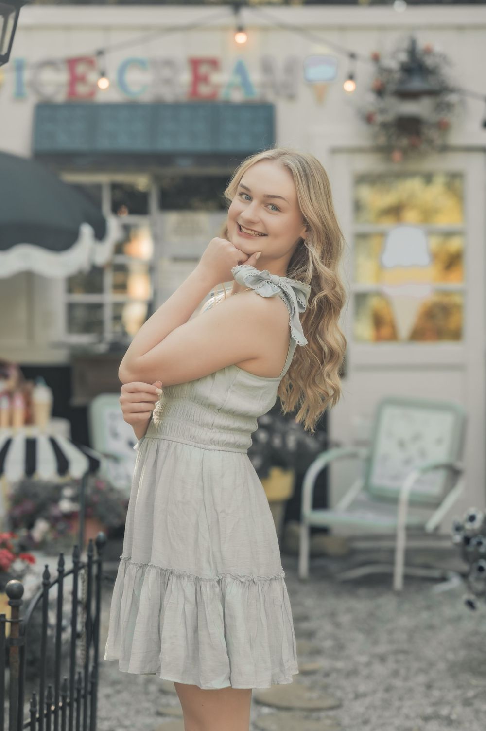 A person in a light gray summer dress poses cheerfully outside an ice cream shop with vintage decor.