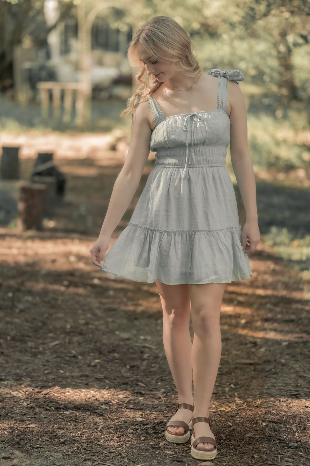 A dreamy summer portrait shows someone in a light gray sundress and sandals standing on a sunlit forest path.