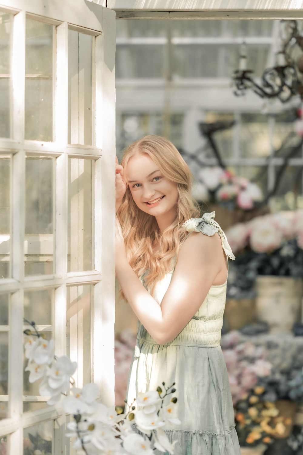 A romantic portrait of a person in a light dress standing by a window in a botanical setting with soft natural lighting.