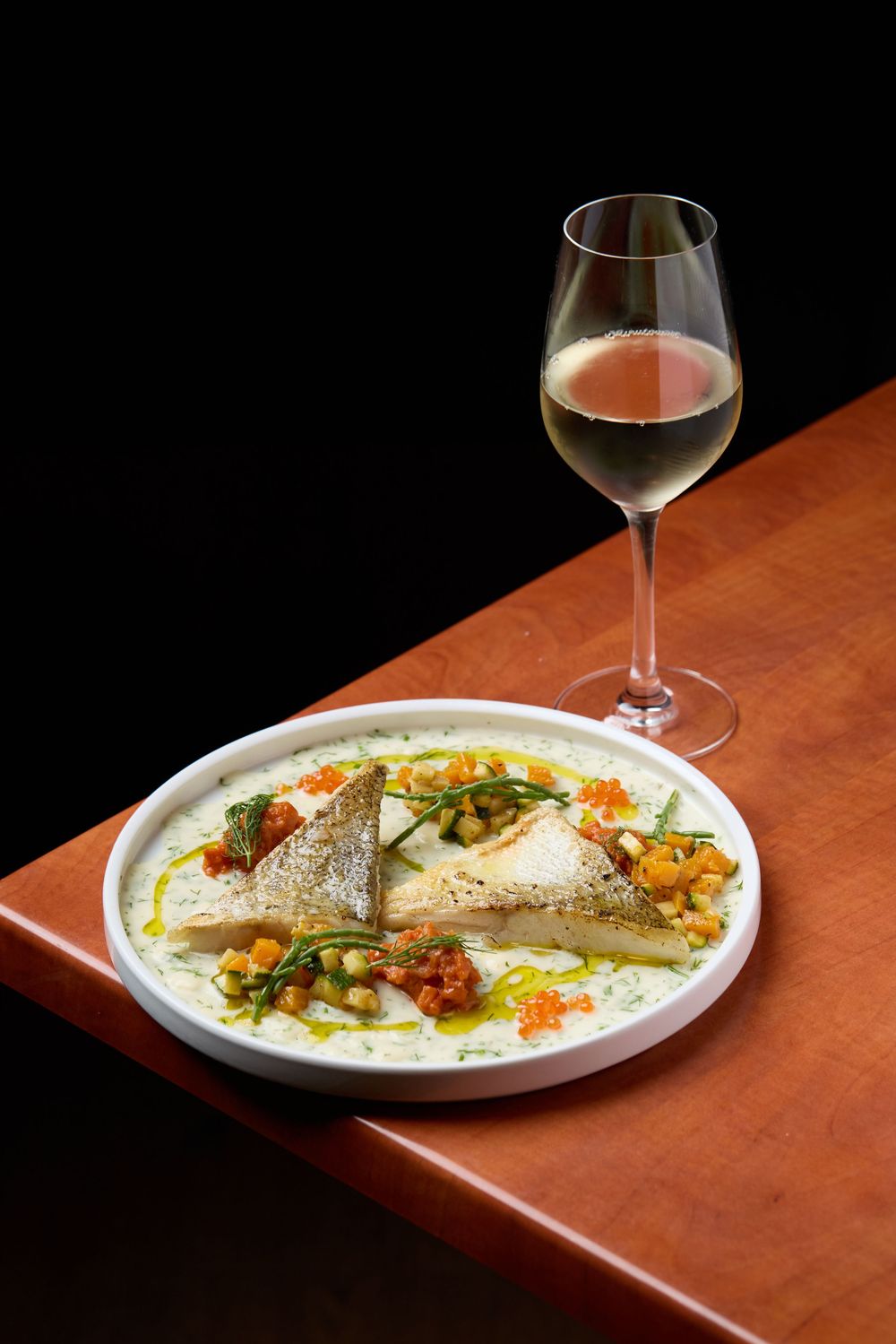 Elegant plated fish dish with wine glass on wooden table against black background.