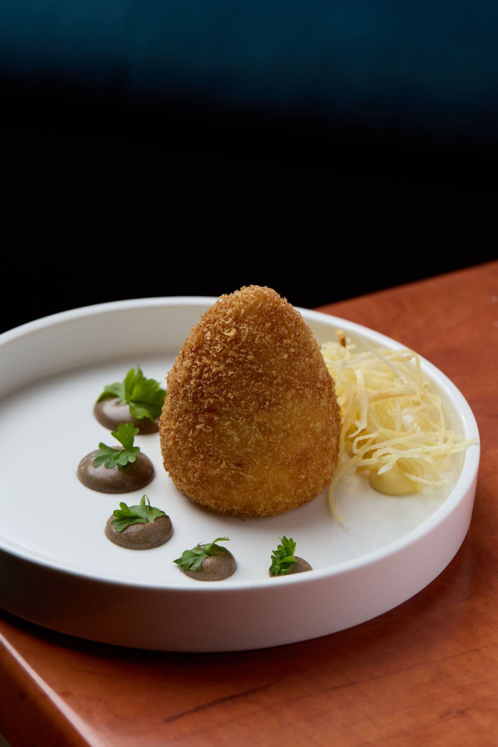 Breaded croquette served with lemon and sauce on white plate against dark background.