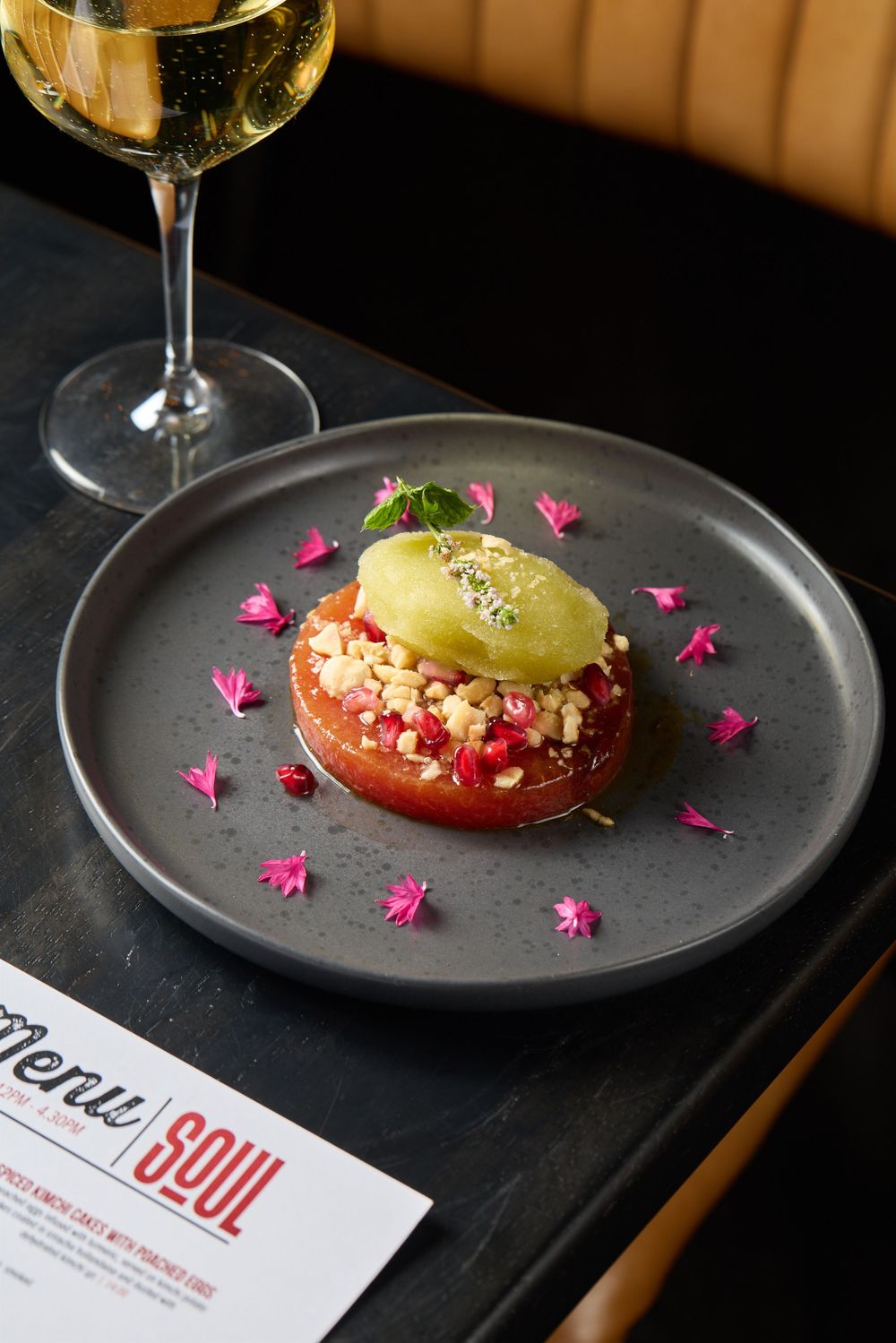 Elegantly plated dessert with pink flower petals and a glass of white wine on a dark plate.