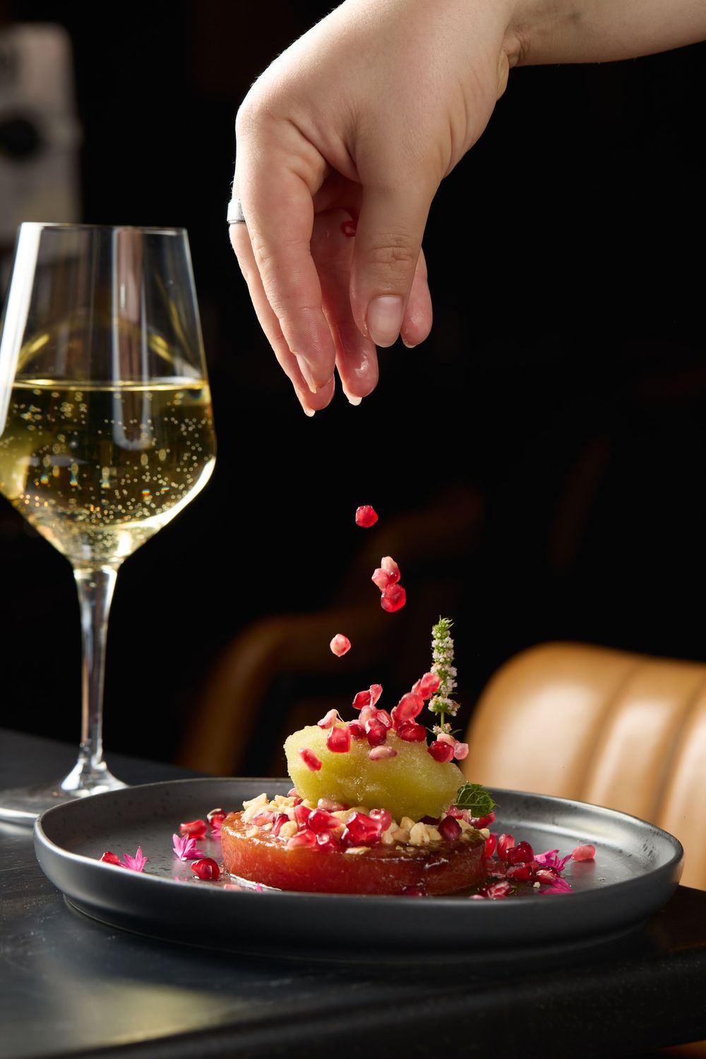 Hands garnishing a plated dessert with pink flower petals and white wine.