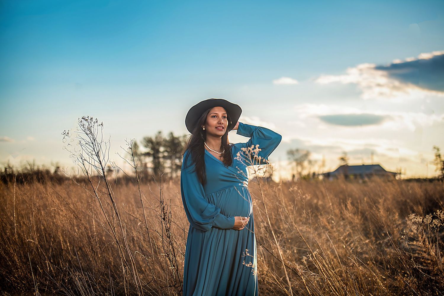 A woman in a blue dress and hat stands in a golden wheat field at sunset.