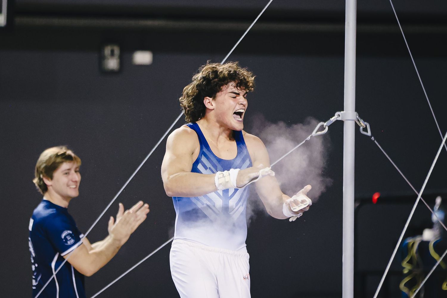 Male gymnast in blue and white uniform performs on high bar apparatus during competition with chalk dust visible.