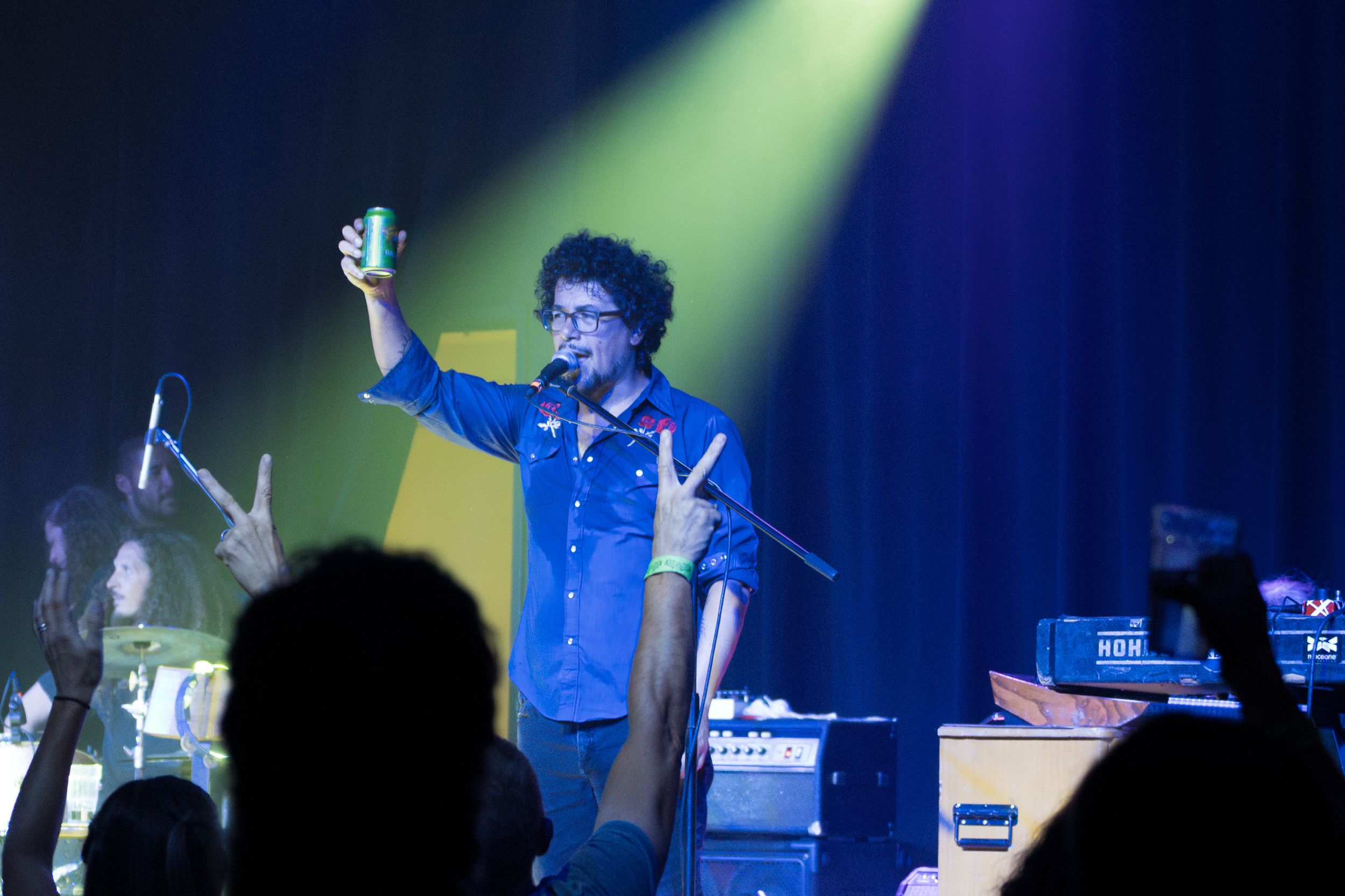A performer in a blue shirt raises their arms enthusiastically on stage during a concert performance with dramatic lighting.