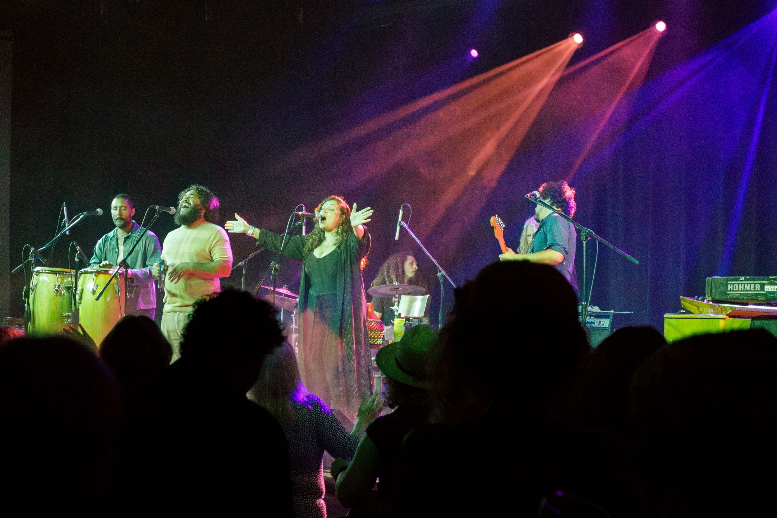 Musicians perform on stage with colorful blue and red lighting effects while silhouetted audience members watch from below.
