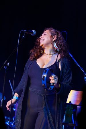 Singer performing on stage in a black outfit laughs joyfully while singing into microphone during live concert performance.