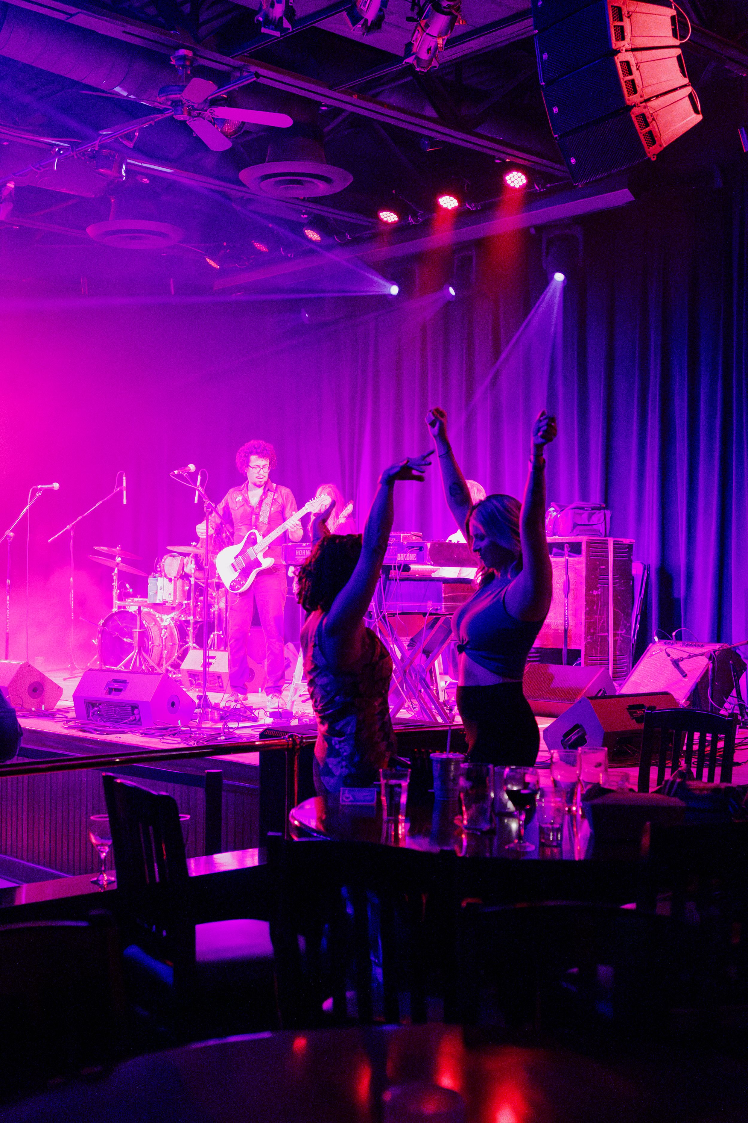 Music concert scene with silhouettes dancing in front of purple and pink stage lights while band performs live.