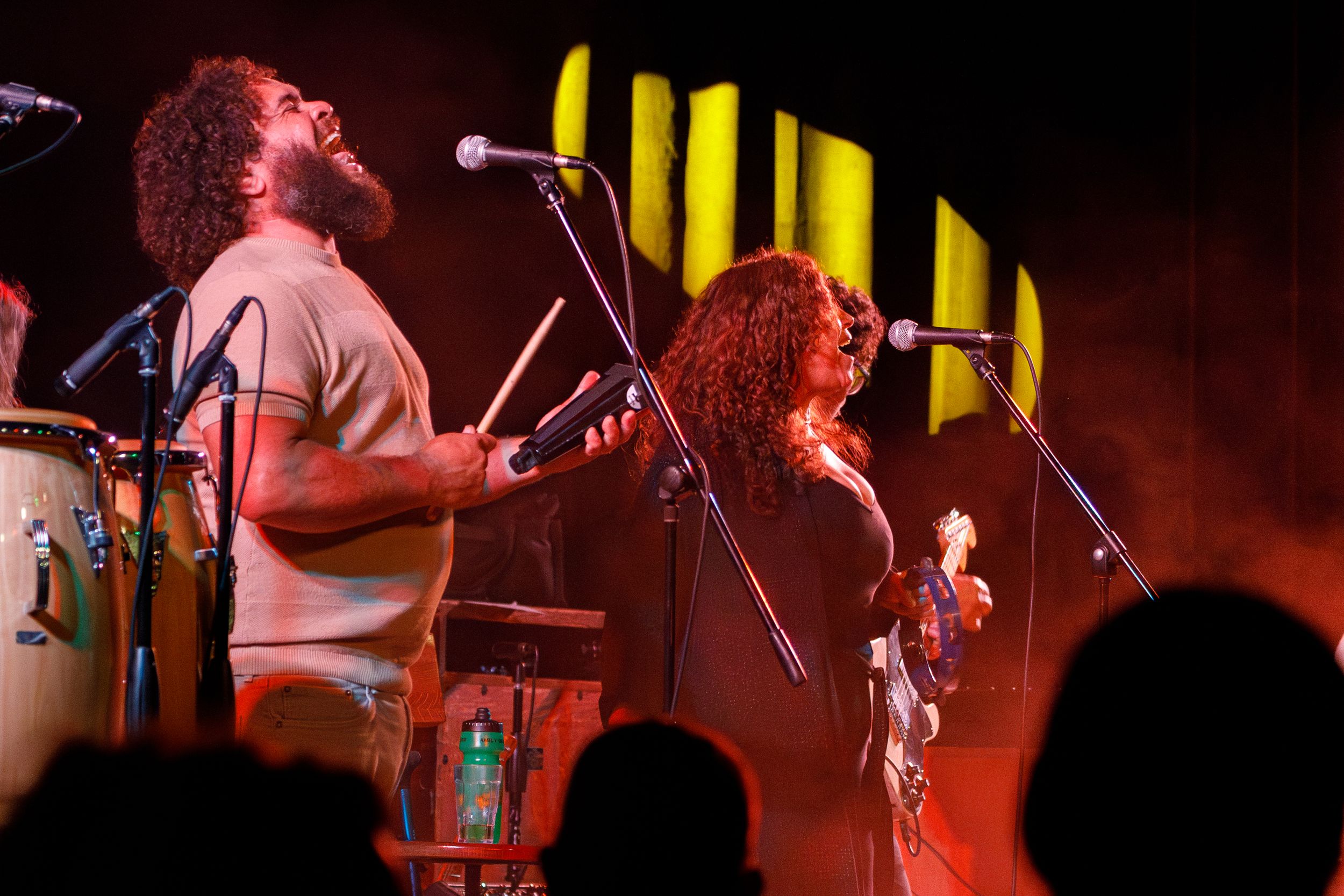 Musicians perform energetically on stage with dramatic red lighting and yellow striped backdrop during a live concert.