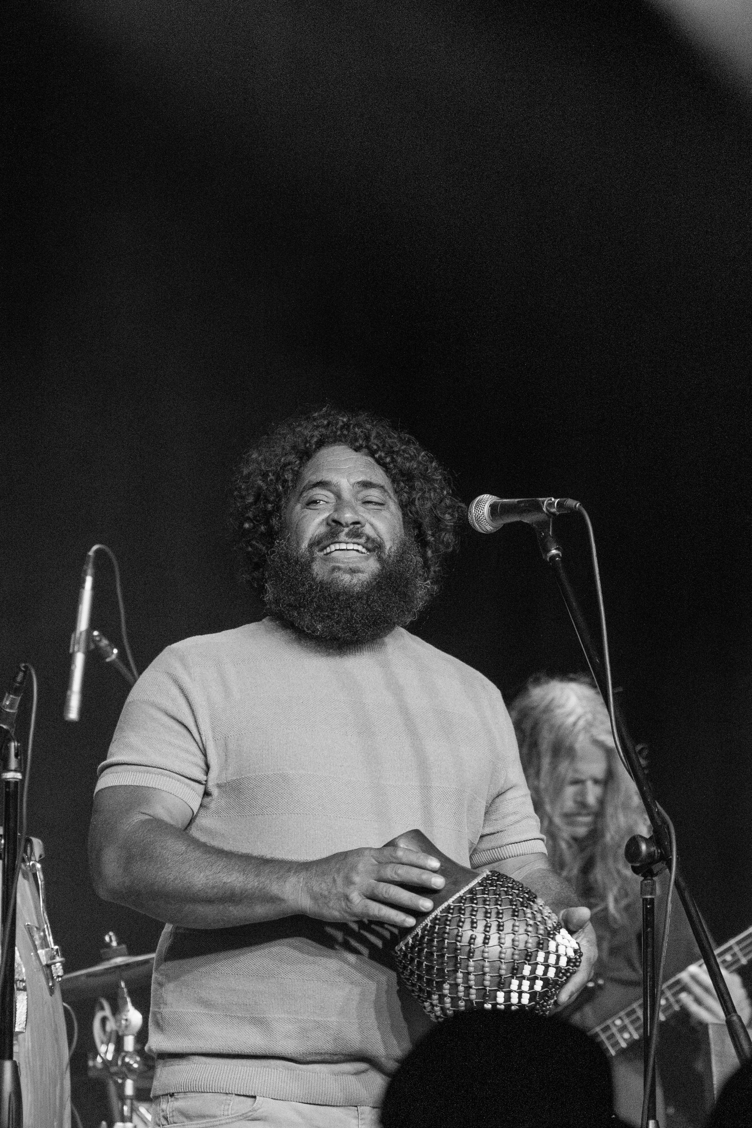 A musician performs on stage with a beard and curly hair, holding a shiny tambourine near microphones in black and white.