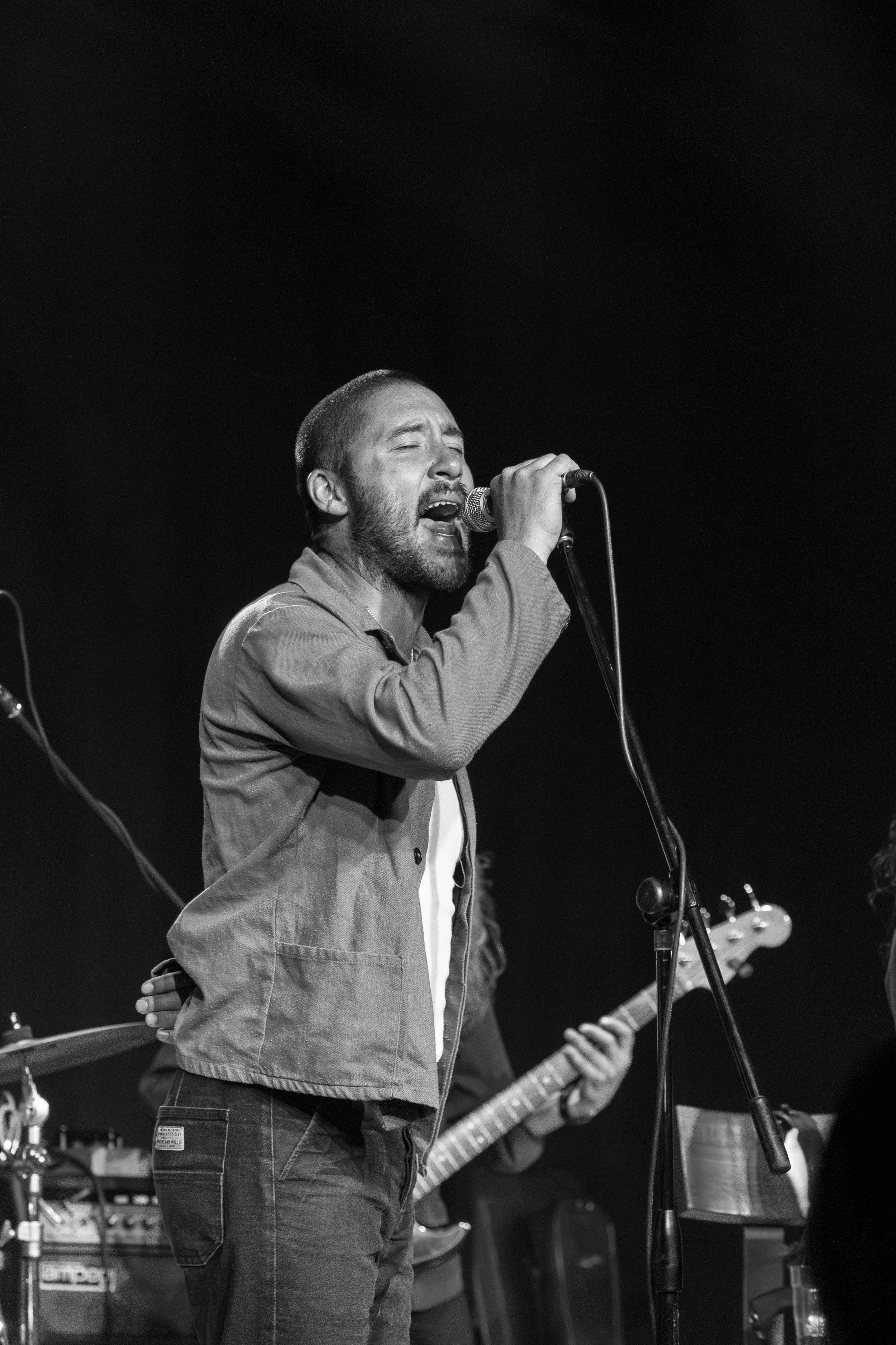 A passionate vocalist sings emotionally into a microphone during a live performance with a band in black and white.