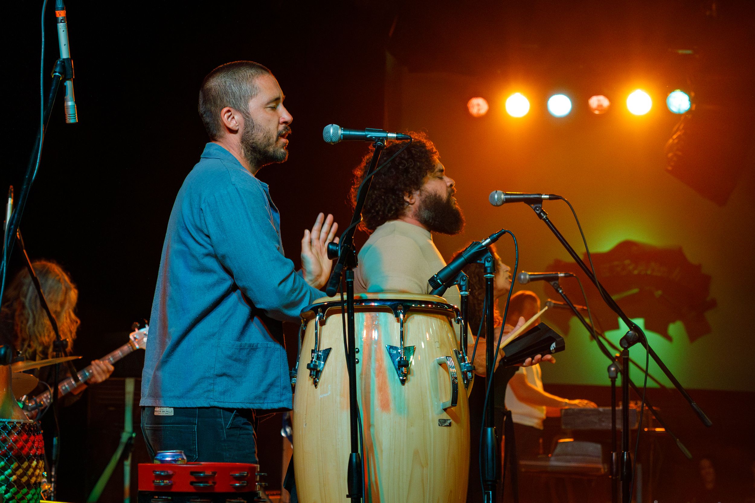 Musicians perform on stage with percussion instruments and microphones under warm orange stage lighting.