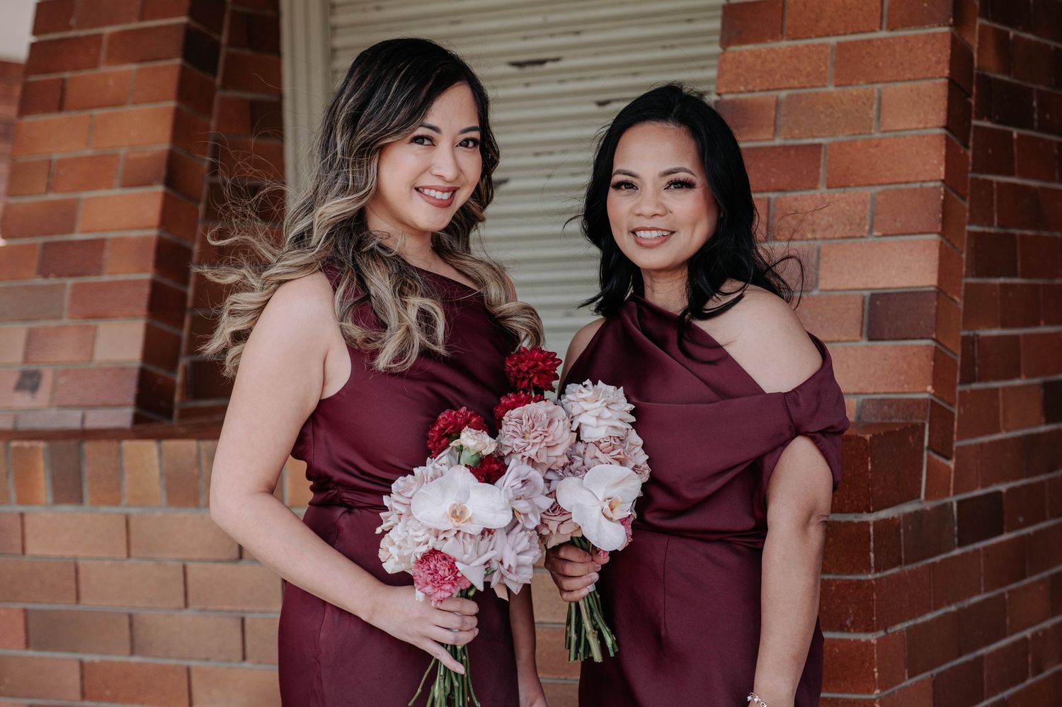 A bride in an elegant white lace gown holds a pink bouquet while standing against a rustic brick wall interior.