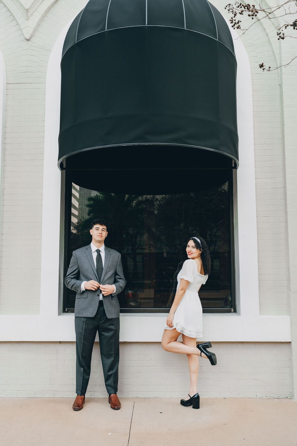A stylish couple poses playfully under a black awning against a white building exterior.