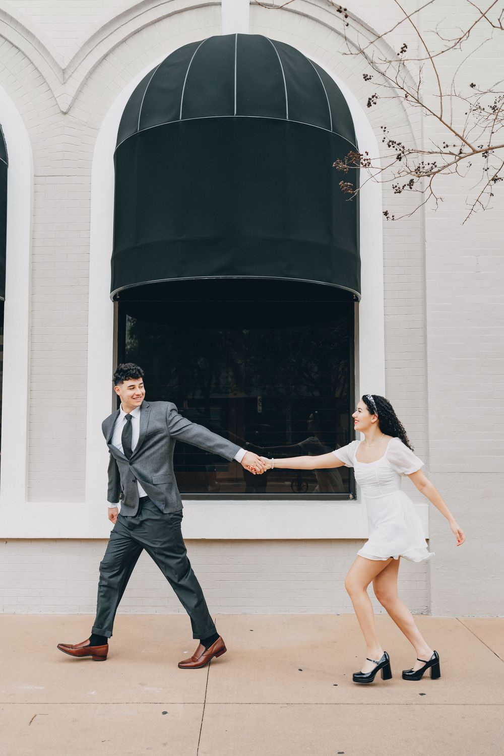 A couple in formal attire playfully dancing outside a building with arched windows and dark awnings.