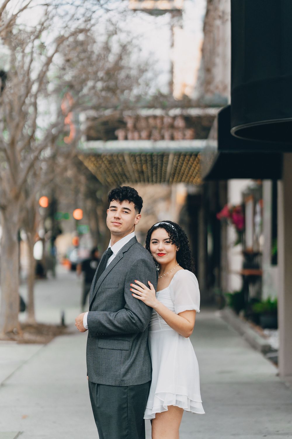 A couple in formal attire poses together on an urban sidewalk with storefronts and trees in the background.