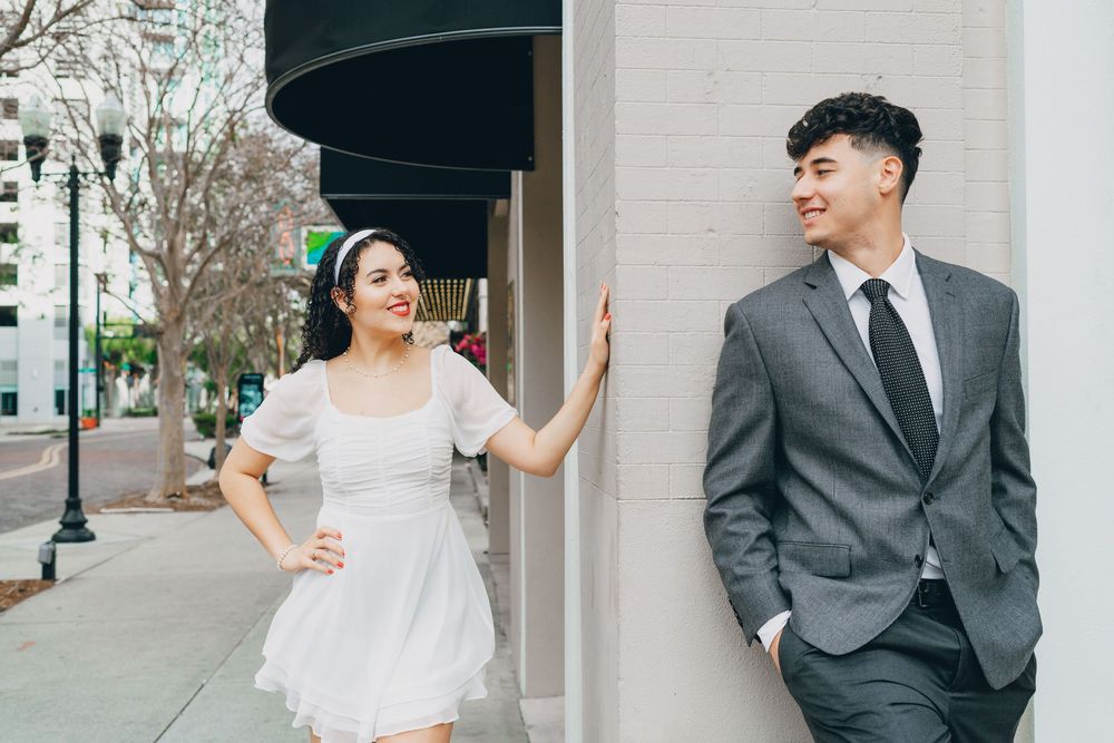 A laughing couple in formal attire pose playfully against a building wall on an urban street.
