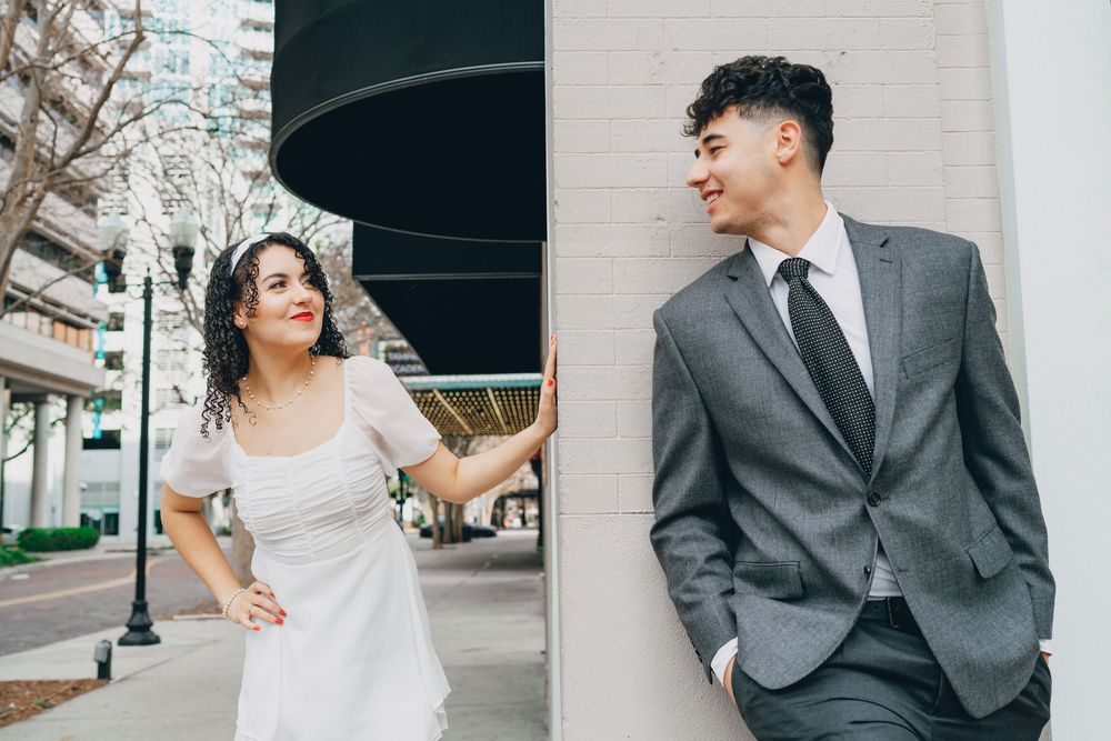 A playful couple in formal attire poses against an urban building exterior.