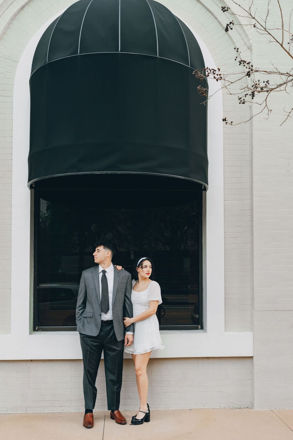 A couple poses beneath a black awning against a white building in formal attire.