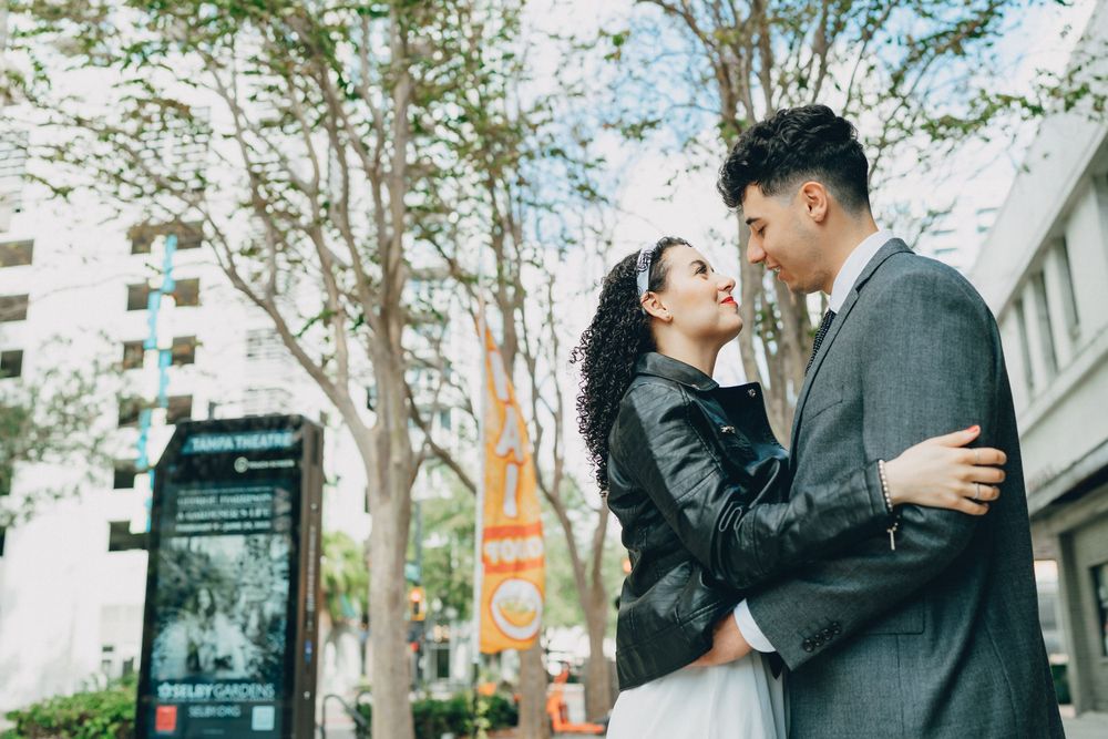 A romantic couple in formal wear embraces while standing on a city street with tall buildings visible behind them.