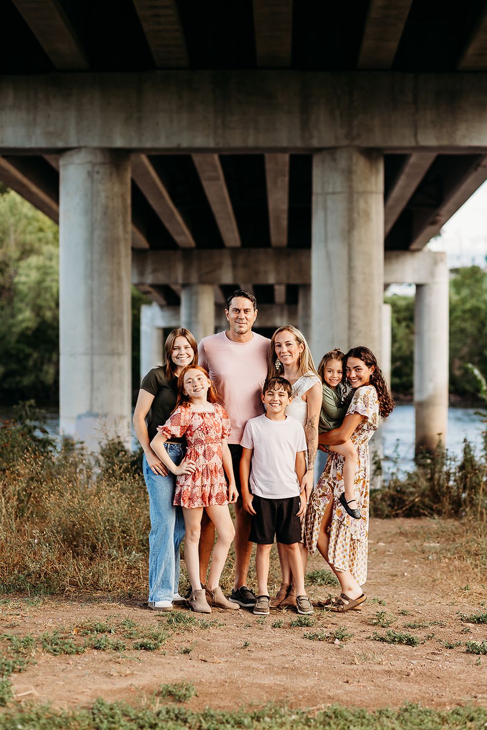 Group poses together for a casual outdoor family portrait under a large concrete bridge on a sunny day.