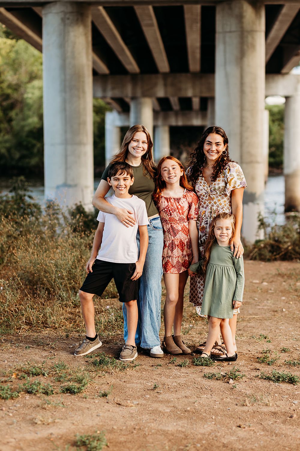 Family group poses together under concrete bridge supports at sunset on dirt ground during outdoor photoshoot.
