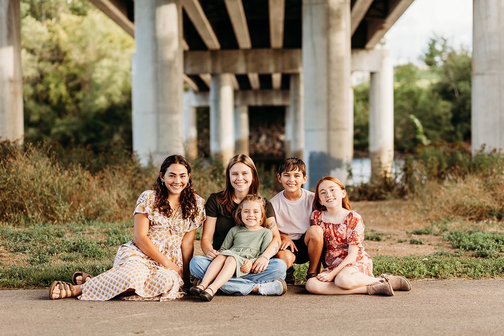 A group of friends sits together in casual outfits under a bridge structure during sunset.
