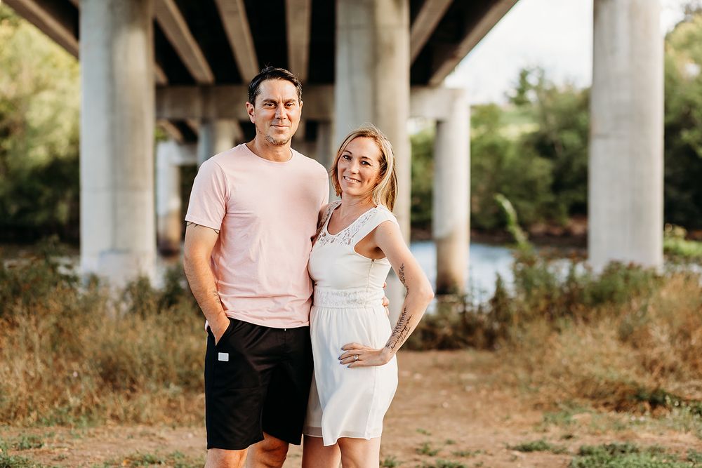 A couple poses outdoors under a concrete bridge on a sunny day, with dry grass and greenery in the background.