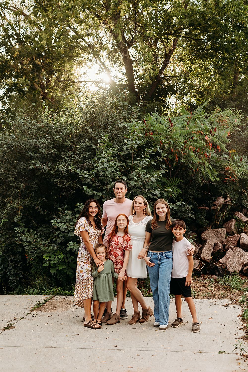 A group of friends pose casually together outdoors on a dirt path surrounded by lush green foliage.