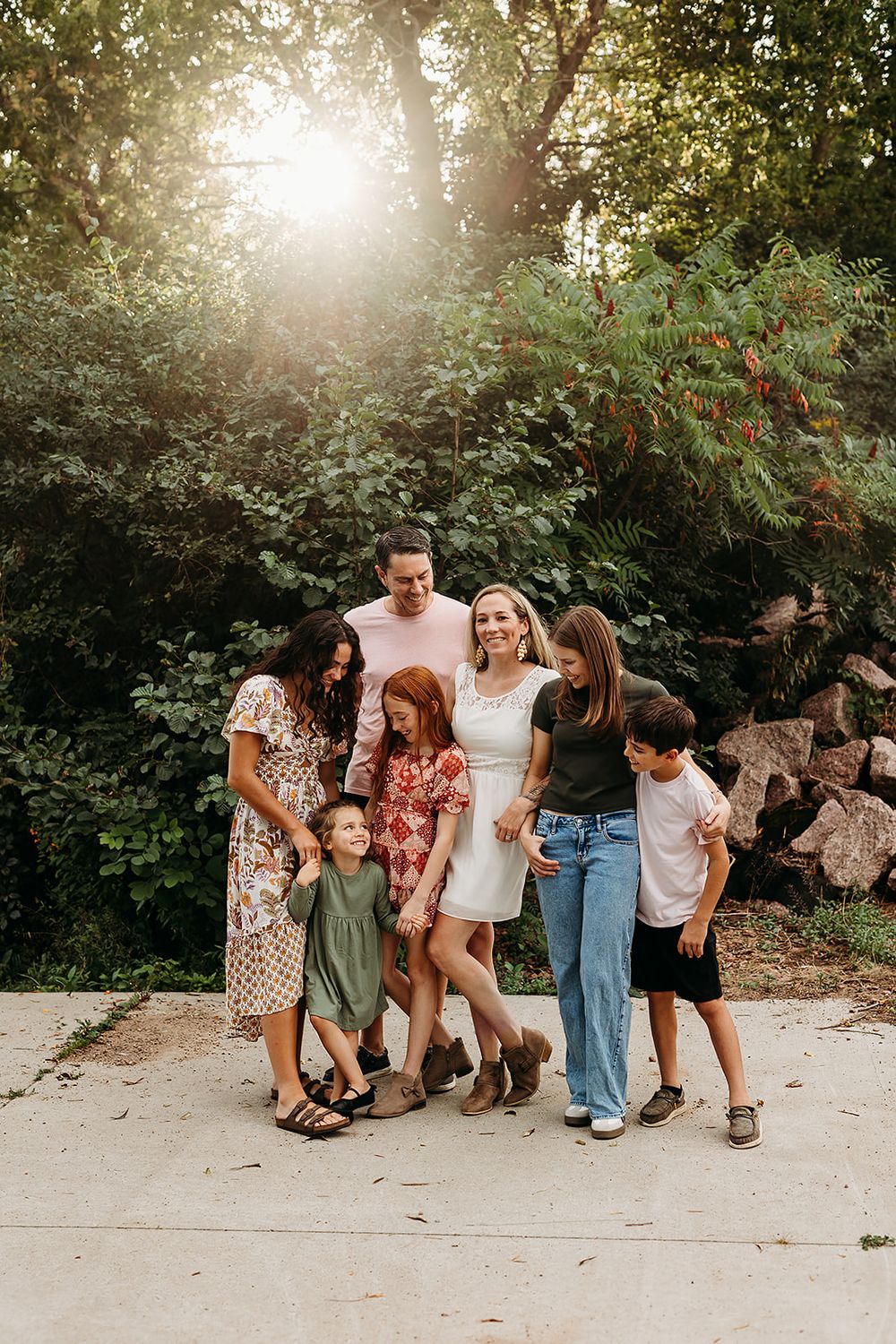 A group stands together outdoors on a path with a brown dog while sunlight filters through trees in the background.