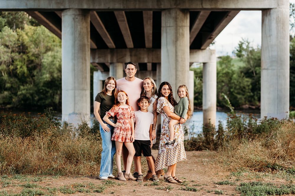 A group stands together under a concrete bridge overpass surrounded by tall grass and natural scenery.