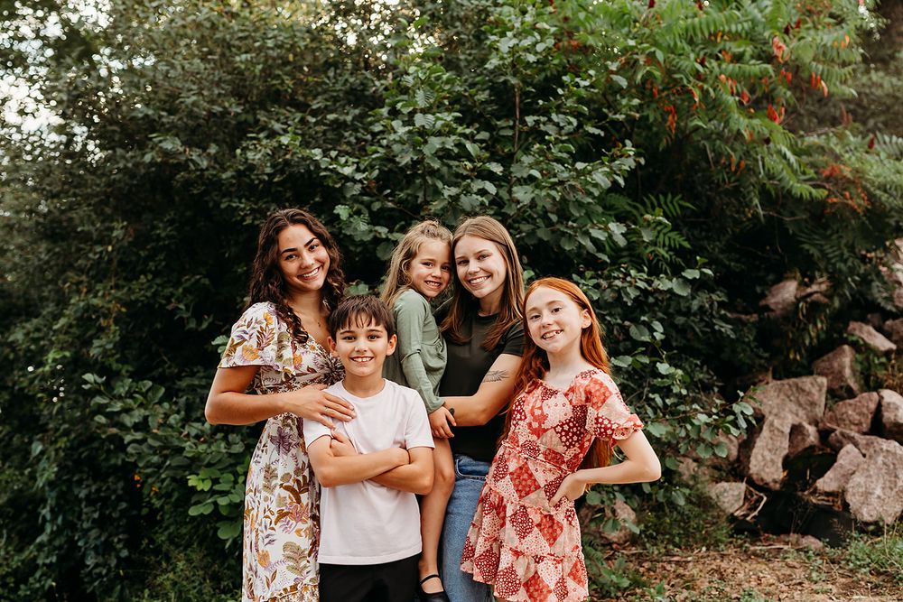 A group portrait in a garden setting with floral dresses and casual attire against lush greenery backdrop.