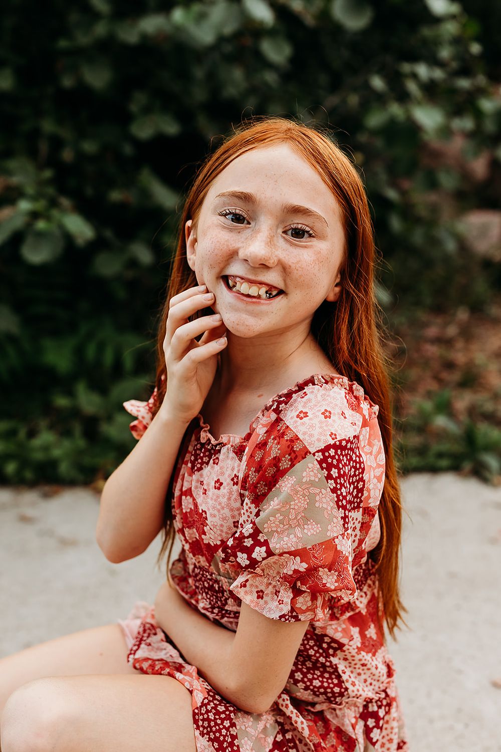 Young person in a red floral dress sits outdoors with a bright smile in a natural garden setting.