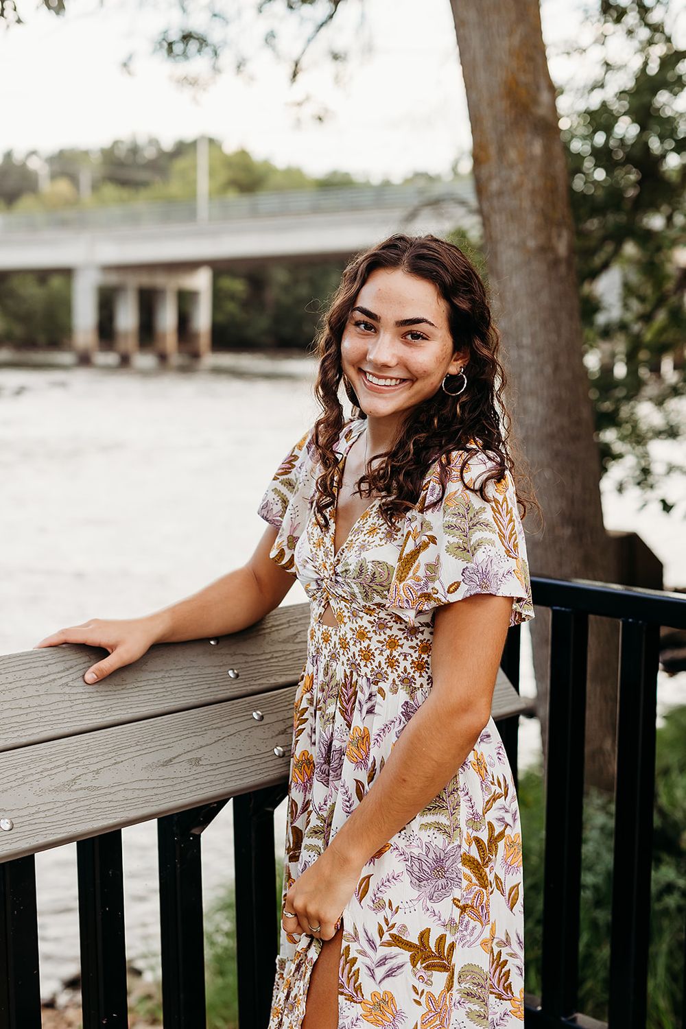 A smiling person in a floral print dress leans against a wooden railing on an outdoor deck during golden hour.